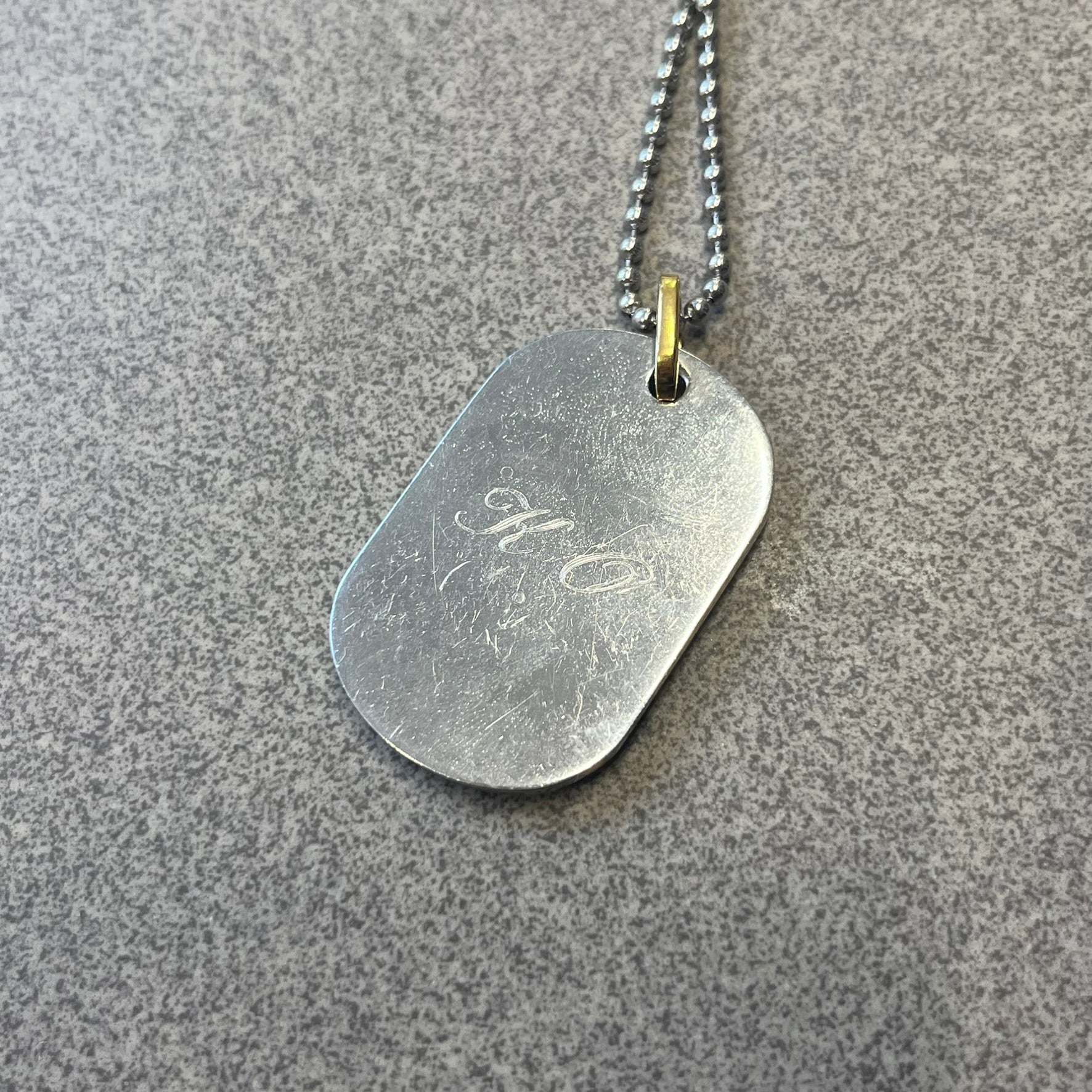 Dolce & Gabbana DJ0637 Gold Heart Necklace features a silver-tone stainless steel ball chain with a dog tag-style pendant. The pendant is engraved with a subtle "I" above a gold-tone heart secured by three visible screws, with "D&G" engraved below to form the "I ❤️ D&G" design. The back of the pendant has a "K.O" engraving from a previous owner. Comes with the original dust bag.
