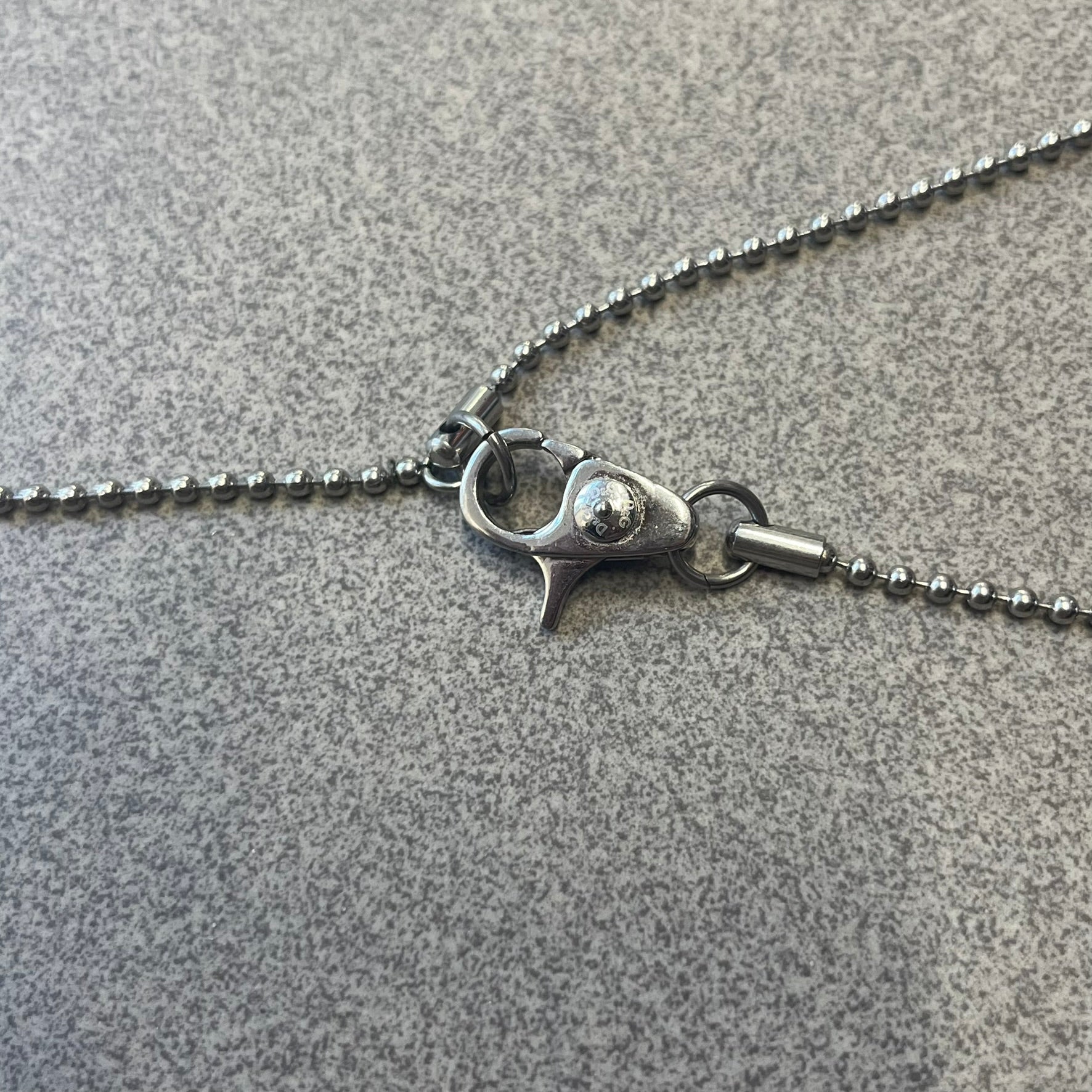 Dolce & Gabbana DJ0637 Gold Heart Necklace features a silver-tone stainless steel ball chain with a dog tag-style pendant. The pendant is engraved with a subtle "I" above a gold-tone heart secured by three visible screws, with "D&G" engraved below to form the "I ❤️ D&G" design. The back of the pendant has a "K.O" engraving from a previous owner. Comes with the original dust bag.
