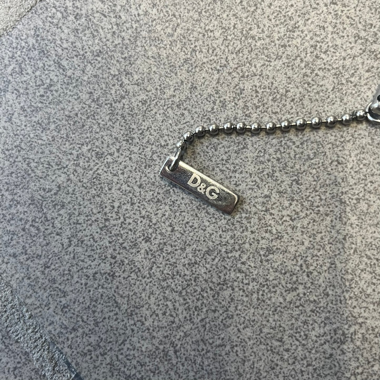 Dolce & Gabbana DJ0637 Gold Heart Necklace features a silver-tone stainless steel ball chain with a dog tag-style pendant. The pendant is engraved with a subtle "I" above a gold-tone heart secured by three visible screws, with "D&G" engraved below to form the "I ❤️ D&G" design. The back of the pendant has a "K.O" engraving from a previous owner. Comes with the original dust bag.
