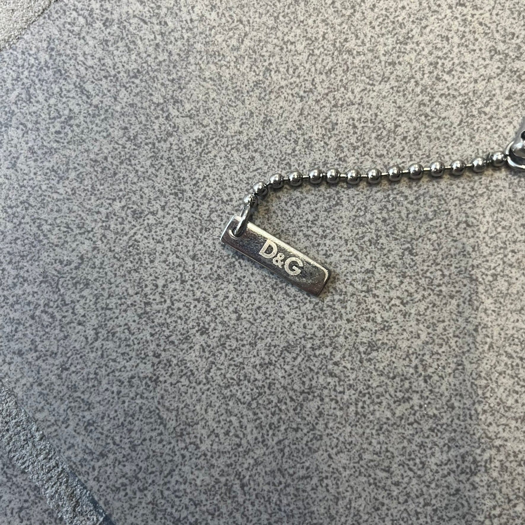 Dolce & Gabbana DJ0637 Gold Heart Necklace features a silver-tone stainless steel ball chain with a dog tag-style pendant. The pendant is engraved with a subtle "I" above a gold-tone heart secured by three visible screws, with "D&G" engraved below to form the "I ❤️ D&G" design. The back of the pendant has a "K.O" engraving from a previous owner. Comes with the original dust bag.
