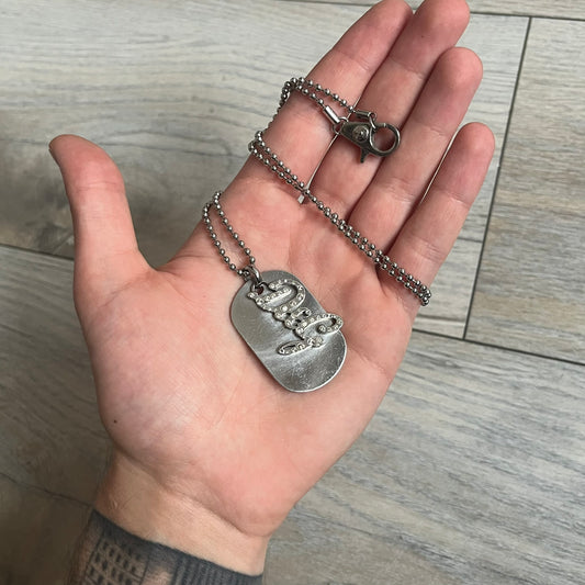 The Dolce &amp; Gabbana Crystal Logo Dog Tag Necklace features a silver-tone ball chain with two pendants: a plain dog tag and a cutout D&amp;G logo adorned with crystals. It has a large clasp engraved with D&amp;G and model markings, from the 2000s.