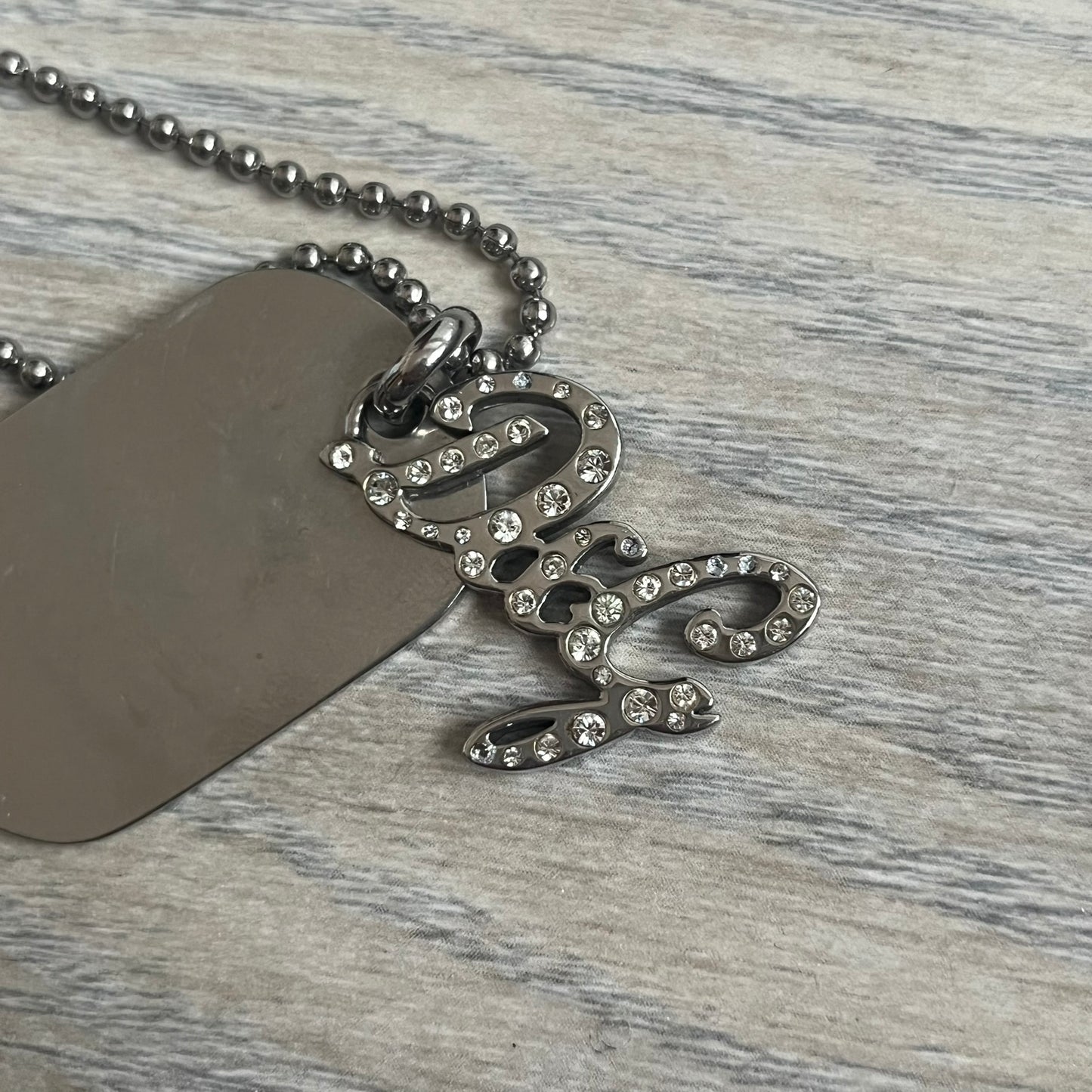 The Dolce &amp; Gabbana Crystal Logo Dog Tag Necklace features a silver-tone ball chain with two pendants: a plain dog tag and a cutout D&amp;G logo adorned with crystals. It has a large clasp engraved with D&amp;G and model markings, from the 2000s.