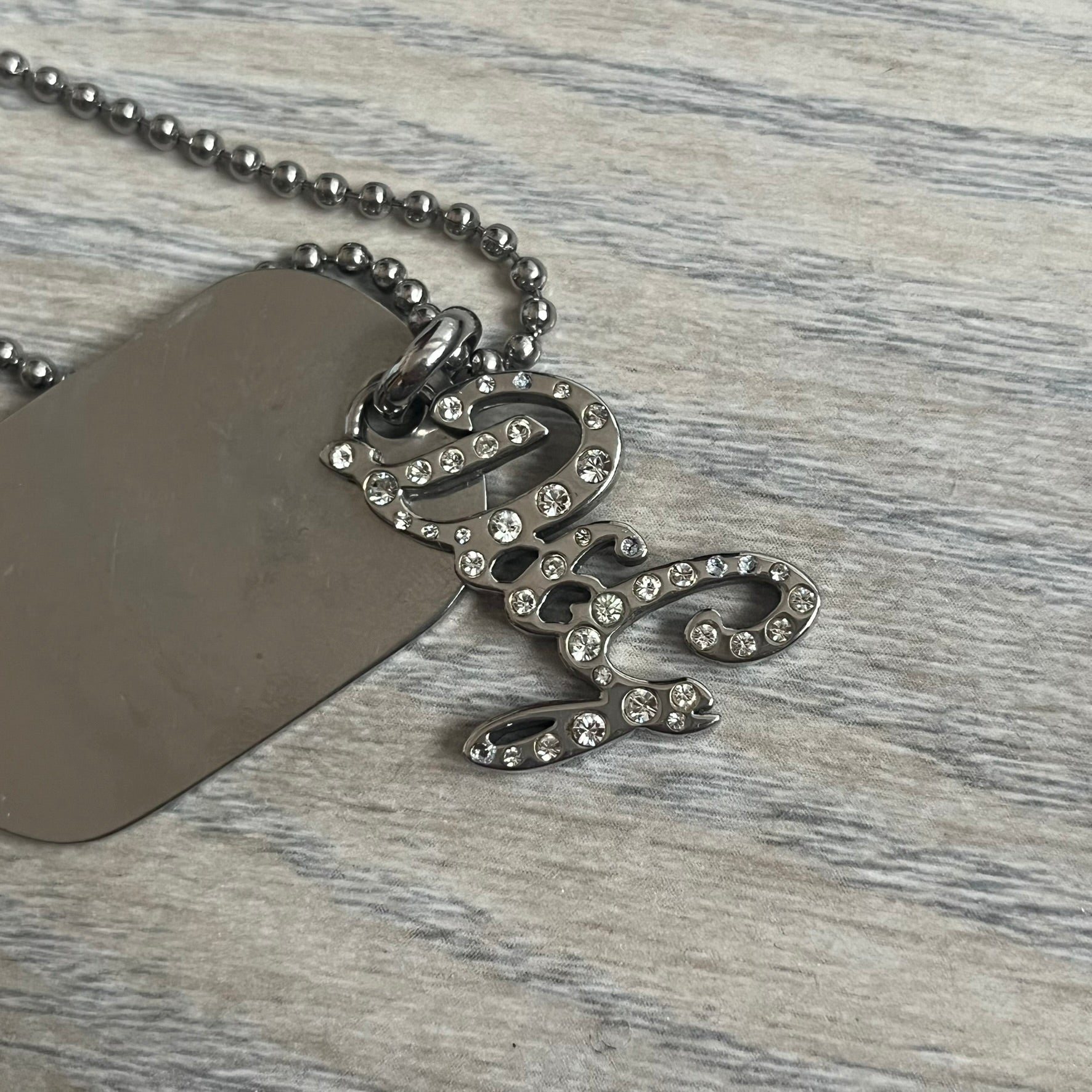 The Dolce &amp; Gabbana Crystal Logo Dog Tag Necklace features a silver-tone ball chain with two pendants: a plain dog tag and a cutout D&amp;G logo adorned with crystals. It has a large clasp engraved with D&amp;G and model markings, from the 2000s.