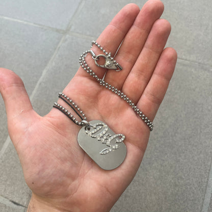 The Dolce &amp; Gabbana Crystal Logo Dog Tag Necklace features a silver-tone ball chain with two pendants: a plain dog tag and a cutout D&amp;G logo adorned with crystals. It has a large clasp engraved with D&amp;G and model markings, from the 2000s.