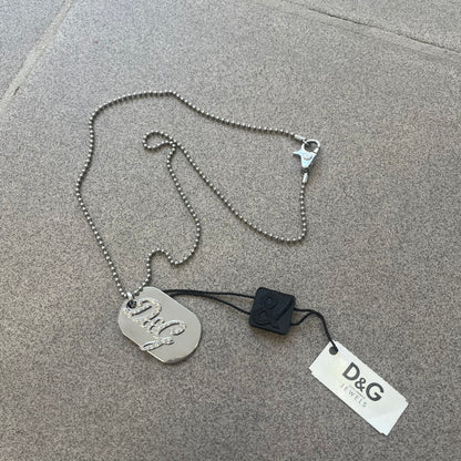 The Dolce &amp; Gabbana Crystal Logo Dog Tag Necklace features a silver-tone ball chain with two pendants: a plain dog tag and a cutout D&amp;G logo adorned with crystals. It has a large clasp engraved with D&amp;G and model markings, from the 2000s.