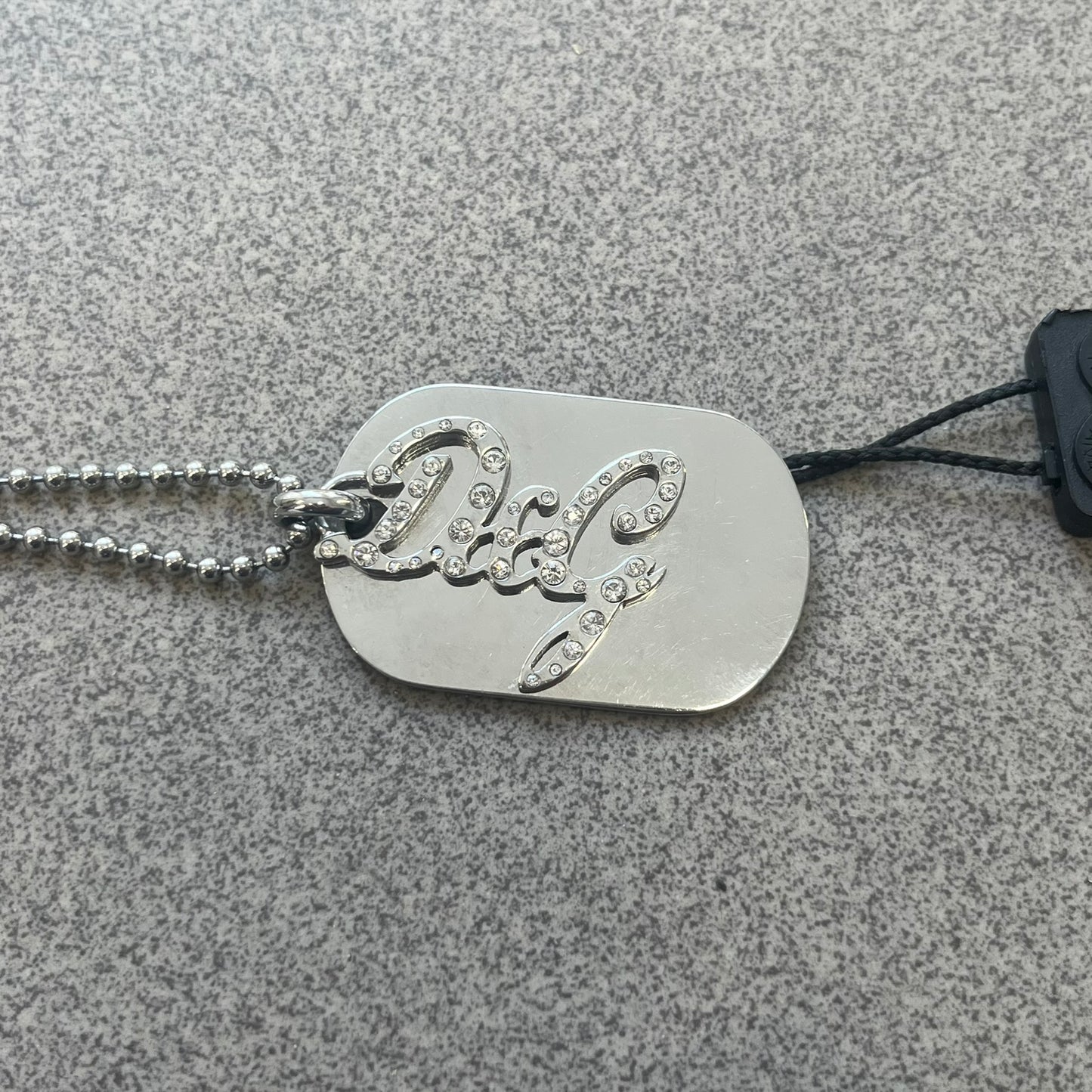 The Dolce &amp; Gabbana Crystal Logo Dog Tag Necklace features a silver-tone ball chain with two pendants: a plain dog tag and a cutout D&amp;G logo adorned with crystals. It has a large clasp engraved with D&amp;G and model markings, from the 2000s.