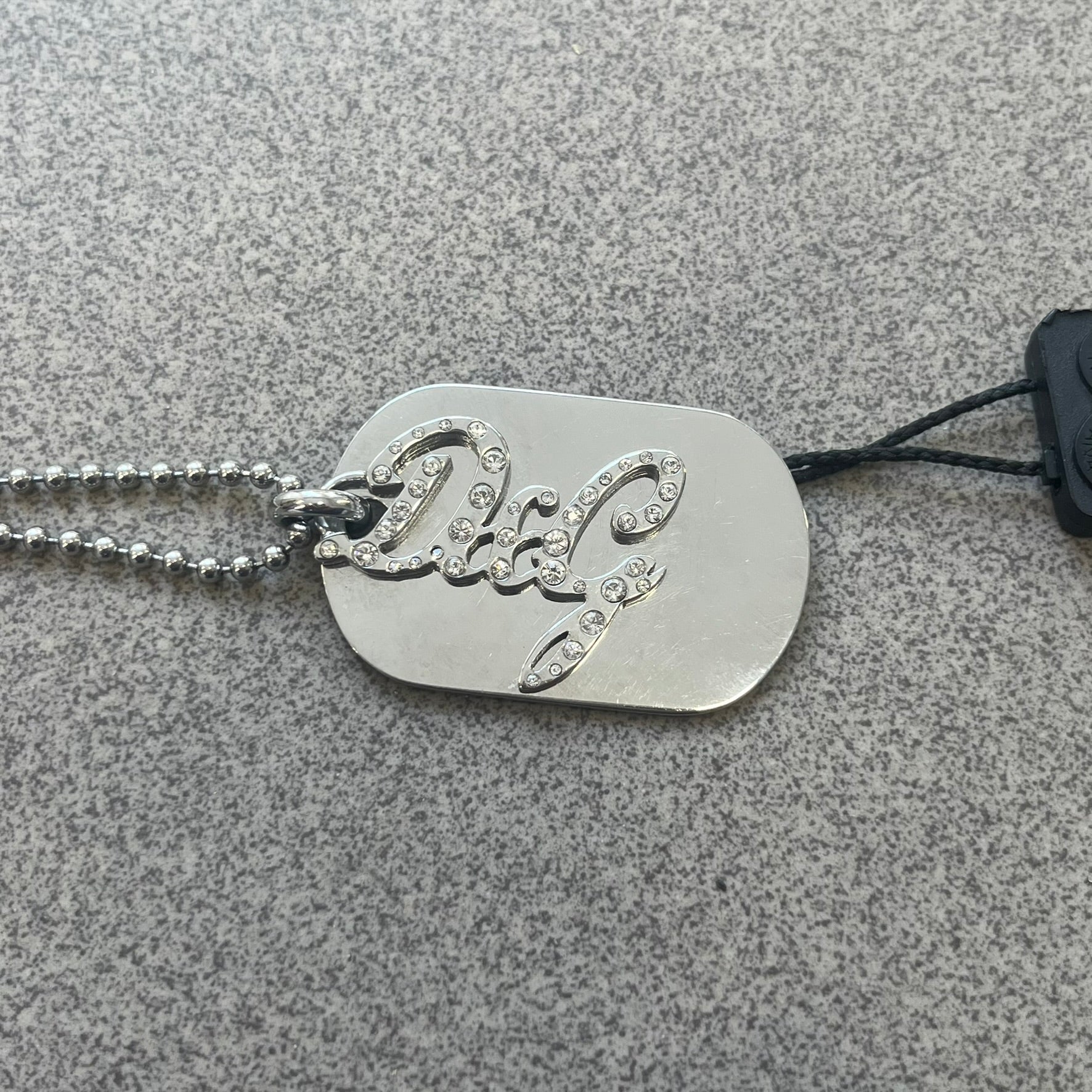 The Dolce &amp; Gabbana Crystal Logo Dog Tag Necklace features a silver-tone ball chain with two pendants: a plain dog tag and a cutout D&amp;G logo adorned with crystals. It has a large clasp engraved with D&amp;G and model markings, from the 2000s.