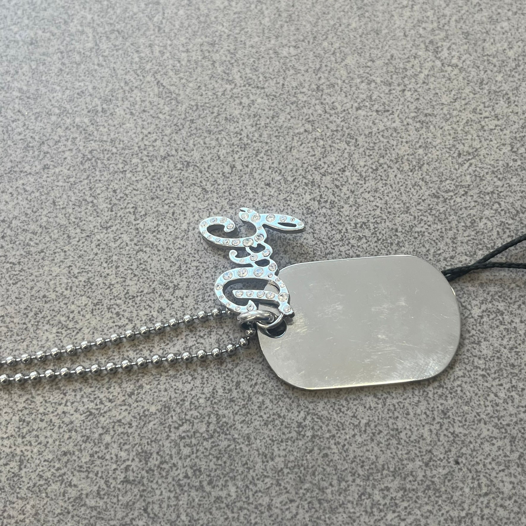 The Dolce &amp; Gabbana Crystal Logo Dog Tag Necklace features a silver-tone ball chain with two pendants: a plain dog tag and a cutout D&amp;G logo adorned with crystals. It has a large clasp engraved with D&amp;G and model markings, from the 2000s.