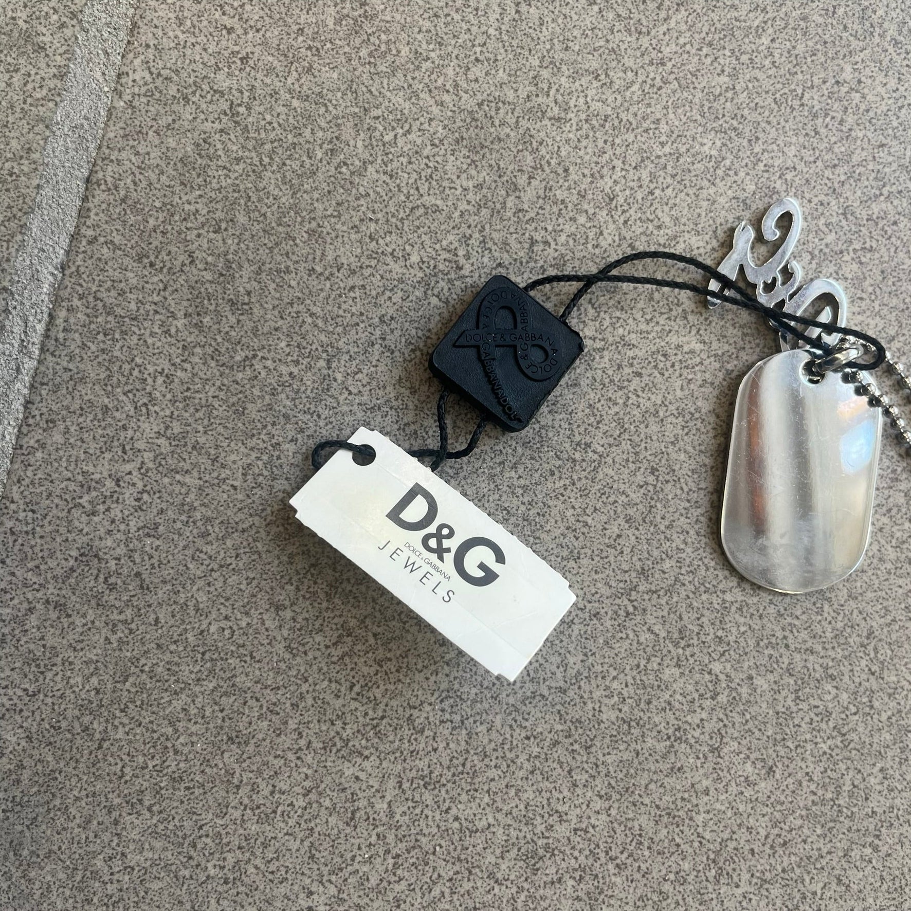 The Dolce &amp; Gabbana Crystal Logo Dog Tag Necklace features a silver-tone ball chain with two pendants: a plain dog tag and a cutout D&amp;G logo adorned with crystals. It has a large clasp engraved with D&amp;G and model markings, from the 2000s.