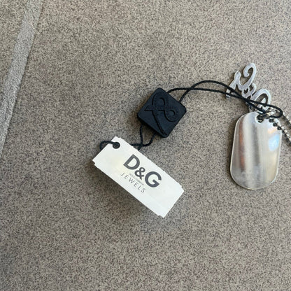 The Dolce &amp; Gabbana Crystal Logo Dog Tag Necklace features a silver-tone ball chain with two pendants: a plain dog tag and a cutout D&amp;G logo adorned with crystals. It has a large clasp engraved with D&amp;G and model markings, from the 2000s.