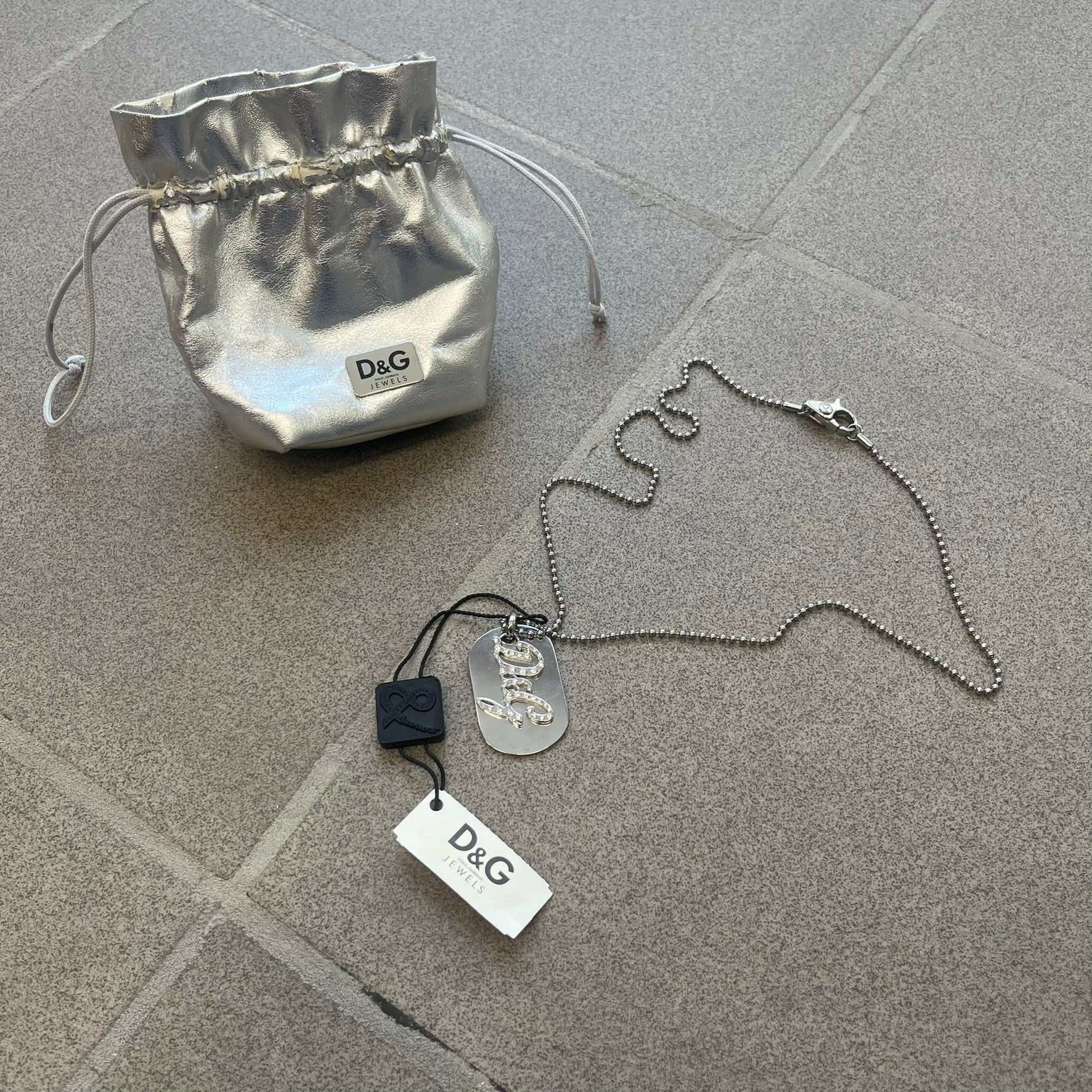 The Dolce &amp; Gabbana Crystal Logo Dog Tag Necklace features a silver-tone ball chain with two pendants: a plain dog tag and a cutout D&amp;G logo adorned with crystals. It has a large clasp engraved with D&amp;G and model markings, from the 2000s.