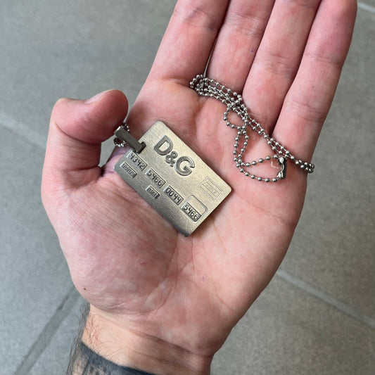 Dolce &amp; Gabbana 2000s Credit Card Necklace (DJ0772) is a rare stainless steel necklace featuring a credit card-shaped pendant. The pendant includes a large raised D&amp;G logo, an engraved D&amp;G logo, and embossed numbers inspired by a real credit card. The chain is not D&amp;G - it's a basic steel ball chain, only the pendant is from Dolce.