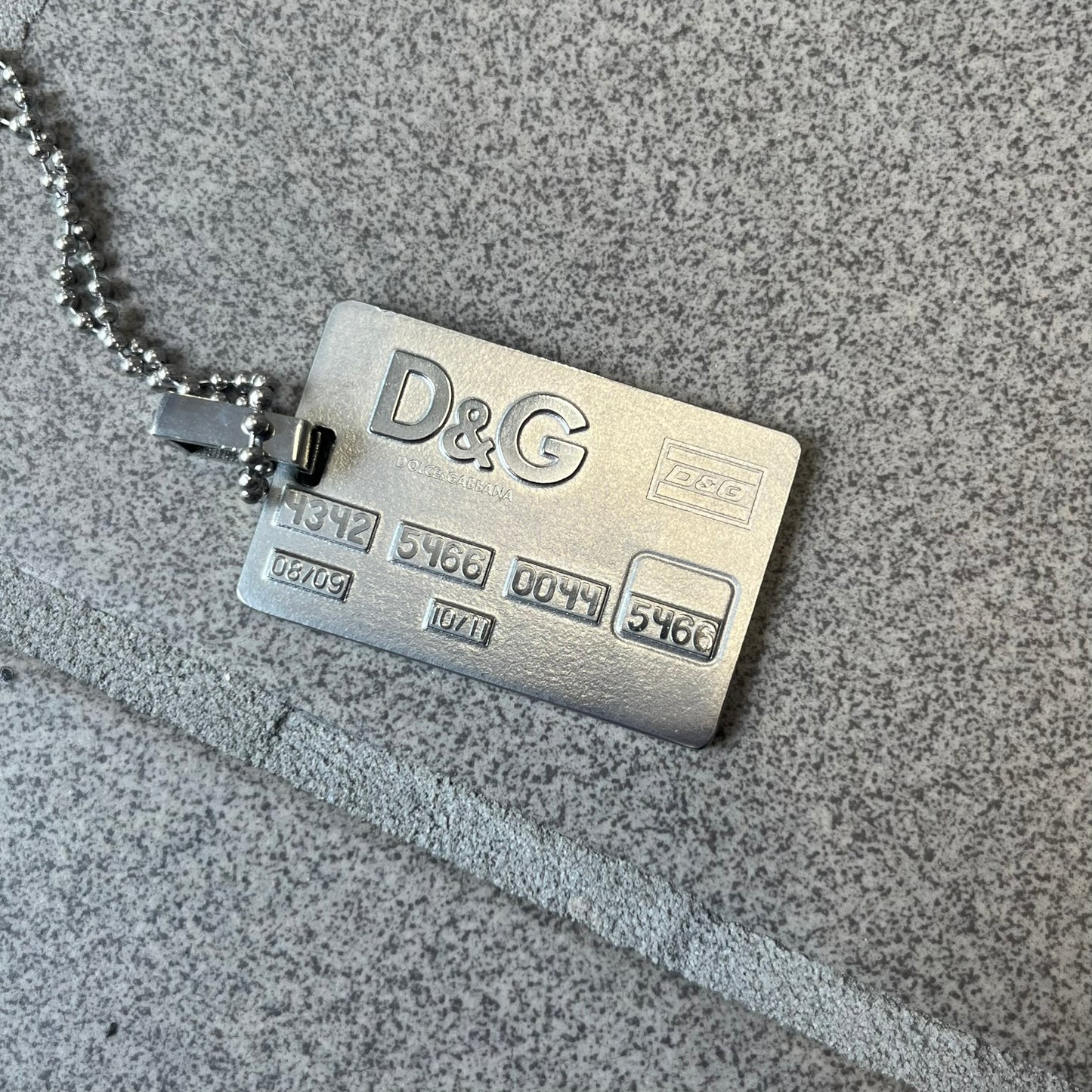 Dolce &amp; Gabbana 2000s Credit Card Necklace (DJ0772) is a rare stainless steel necklace featuring a credit card-shaped pendant. The pendant includes a large raised D&amp;G logo, an engraved D&amp;G logo, and embossed numbers inspired by a real credit card. The chain is not D&amp;G - it's a basic steel ball chain, only the pendant is from Dolce.
