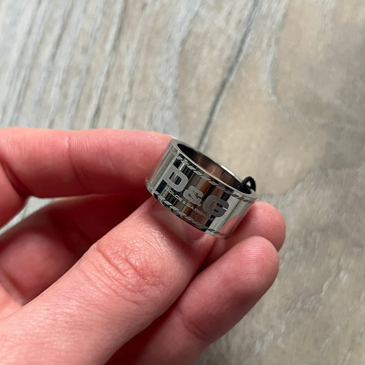 Dolce & Gabbana DJ0836 Logo Ring is a silver-tone stainless steel ring featuring engraved D&G logos in the center. The top and bottom edges have engraved rope-style lines running continuously around the band, giving it a subtle textured detail.
