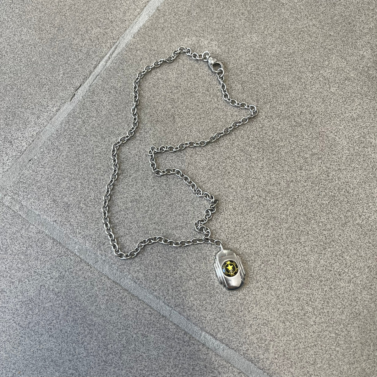 Dolce & Gabbana DJ1067 Gold Star Necklace features a silver-toned rectangular pendant with a black background and a circular plastic center, surrounded by gold D&G logos and a gold star with "1994" text. It comes on a circle-link chain with a large clasp.