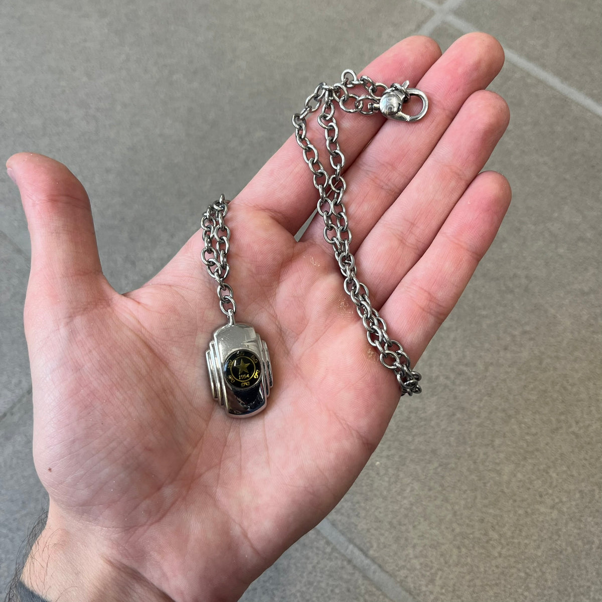 Dolce & Gabbana DJ1067 Gold Star Necklace features a silver-toned rectangular pendant with a black background and a circular plastic center, surrounded by gold D&G logos and a gold star with "1994" text. It comes on a circle-link chain with a large clasp.