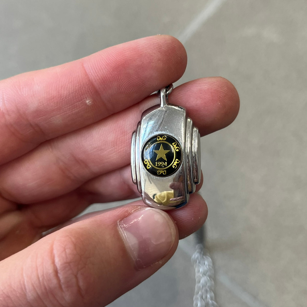 Dolce & Gabbana DJ1067 Gold Star Necklace features a silver-toned rectangular pendant with a black background and a circular plastic center, surrounded by gold D&G logos and a gold star with "1994" text. It comes on a circle-link chain with a large clasp.