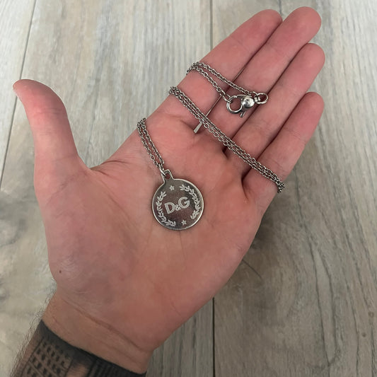 Dolce & Gabbana DJ1087 Medal Necklace is a stainless steel chain necklace featuring a circular 2D pendant. The front design includes an engraved D&G logo surrounded by laurel wreath detailing, with small star accents at the top and bottom. The reverse side of the pendant is plain and smooth. It comes with a thin chain, lobster clasp closure, and a black dust bag.