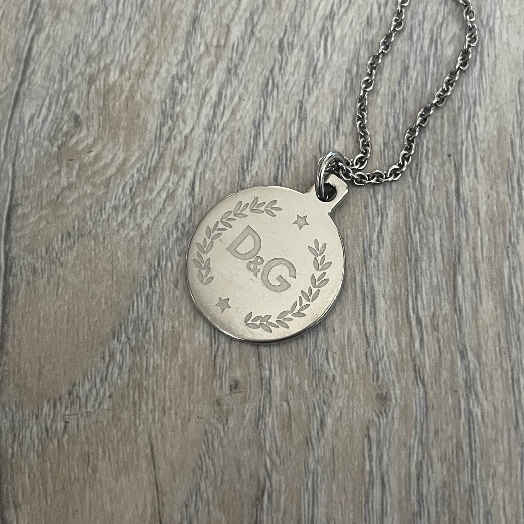 Dolce & Gabbana DJ1087 Medal Necklace is a stainless steel chain necklace featuring a circular 2D pendant. The front design includes an engraved D&G logo surrounded by laurel wreath detailing, with small star accents at the top and bottom. The reverse side of the pendant is plain and smooth. It comes with a thin chain, lobster clasp closure, and a black dust bag.