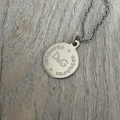 Dolce & Gabbana DJ1087 Medal Necklace is a stainless steel chain necklace featuring a circular 2D pendant. The front design includes an engraved D&G logo surrounded by laurel wreath detailing, with small star accents at the top and bottom. The reverse side of the pendant is plain and smooth. It comes with a thin chain, lobster clasp closure, and a black dust bag.