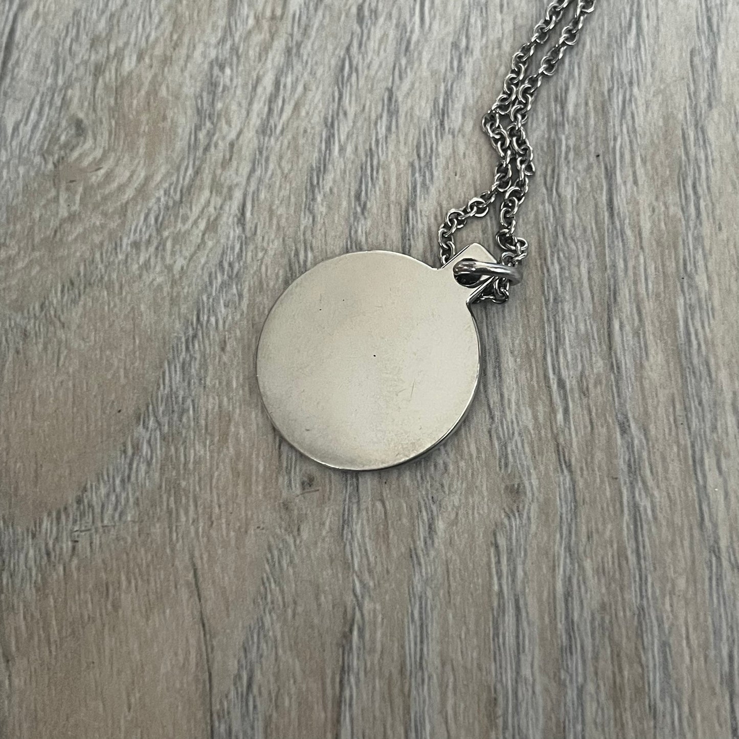 Dolce & Gabbana DJ1087 Medal Necklace is a stainless steel chain necklace featuring a circular 2D pendant. The front design includes an engraved D&G logo surrounded by laurel wreath detailing, with small star accents at the top and bottom. The reverse side of the pendant is plain and smooth. It comes with a thin chain, lobster clasp closure, and a black dust bag.