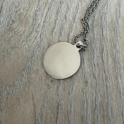 Dolce & Gabbana DJ1087 Medal Necklace is a stainless steel chain necklace featuring a circular 2D pendant. The front design includes an engraved D&G logo surrounded by laurel wreath detailing, with small star accents at the top and bottom. The reverse side of the pendant is plain and smooth. It comes with a thin chain, lobster clasp closure, and a black dust bag.