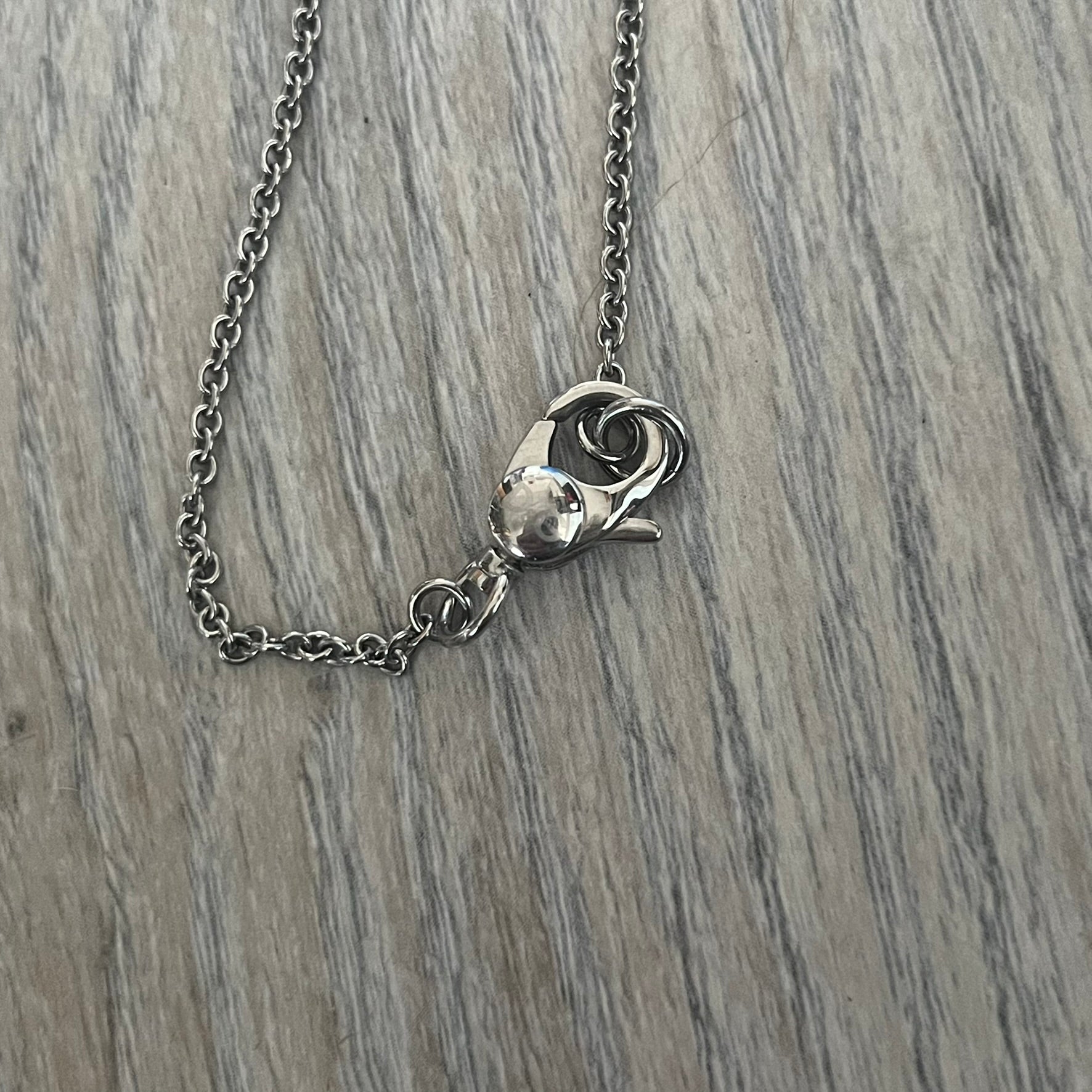 Dolce & Gabbana DJ1087 Medal Necklace is a stainless steel chain necklace featuring a circular 2D pendant. The front design includes an engraved D&G logo surrounded by laurel wreath detailing, with small star accents at the top and bottom. The reverse side of the pendant is plain and smooth. It comes with a thin chain, lobster clasp closure, and a black dust bag.