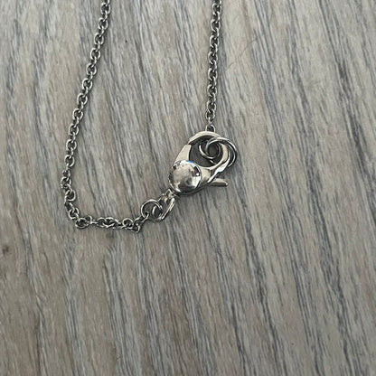 Dolce & Gabbana DJ1087 Medal Necklace is a stainless steel chain necklace featuring a circular 2D pendant. The front design includes an engraved D&G logo surrounded by laurel wreath detailing, with small star accents at the top and bottom. The reverse side of the pendant is plain and smooth. It comes with a thin chain, lobster clasp closure, and a black dust bag.