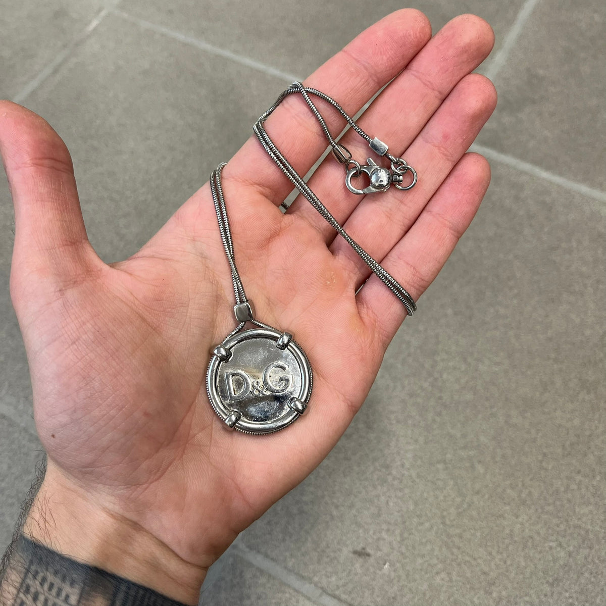 Dolce & Gabbana DJ1115 Circular Necklace is a stainless steel necklace with a circular pendant featuring a raised D&G logo in the center. A thin chain wraps around the pendant outline, held in place by small square clips at four points. It comes with a dust bag.