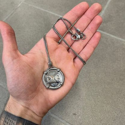 Dolce & Gabbana DJ1115 Circular Necklace is a stainless steel necklace with a circular pendant featuring a raised D&G logo in the center. A thin chain wraps around the pendant outline, held in place by small square clips at four points. It comes with a dust bag.