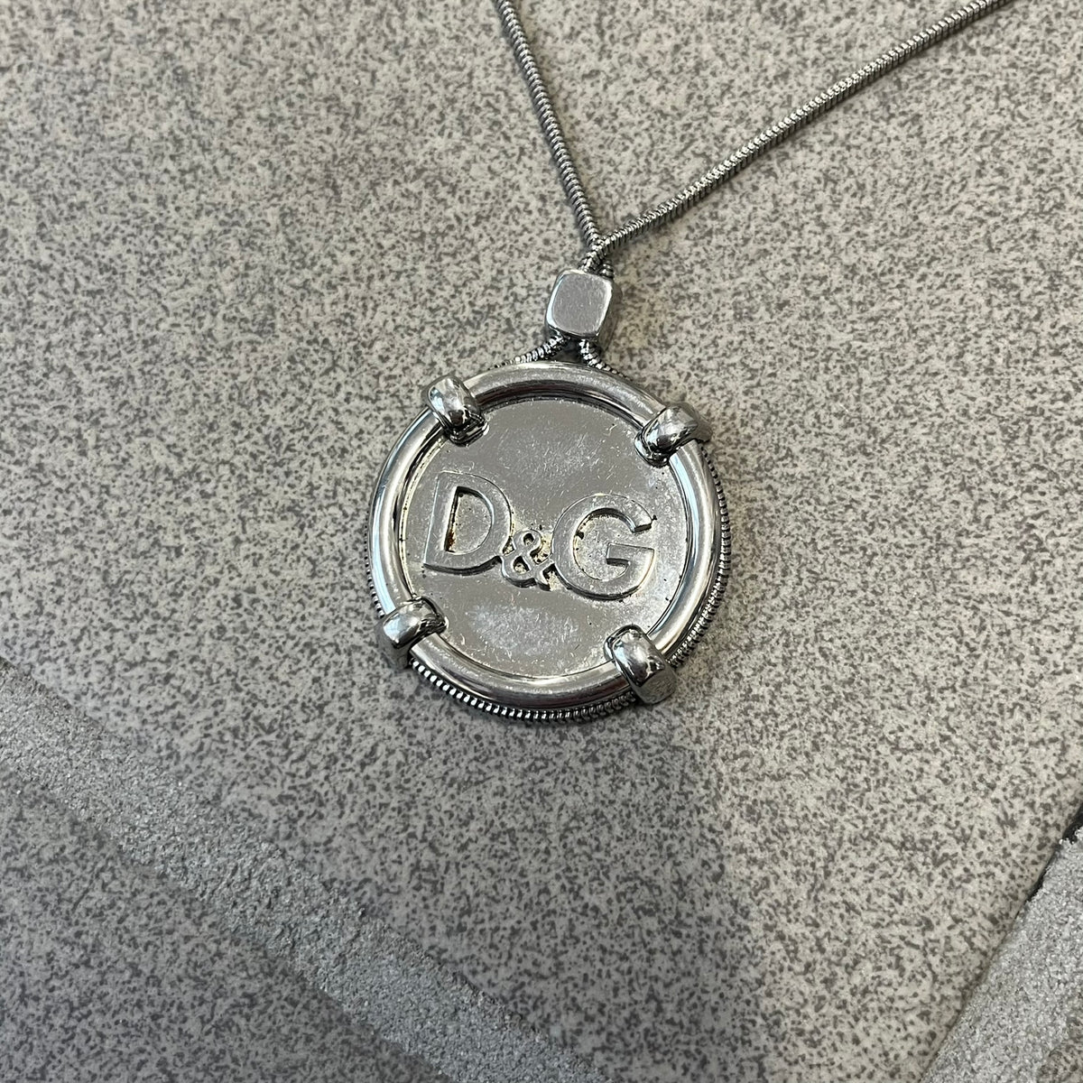 Dolce & Gabbana DJ1115 Circular Necklace is a stainless steel necklace with a circular pendant featuring a raised D&G logo in the center. A thin chain wraps around the pendant outline, held in place by small square clips at four points. It comes with a dust bag.