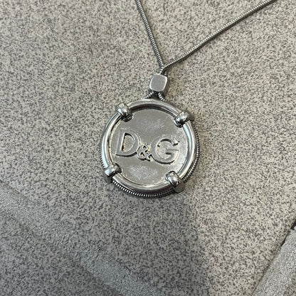 Dolce & Gabbana DJ1115 Circular Necklace is a stainless steel necklace with a circular pendant featuring a raised D&G logo in the center. A thin chain wraps around the pendant outline, held in place by small square clips at four points. It comes with a dust bag.
