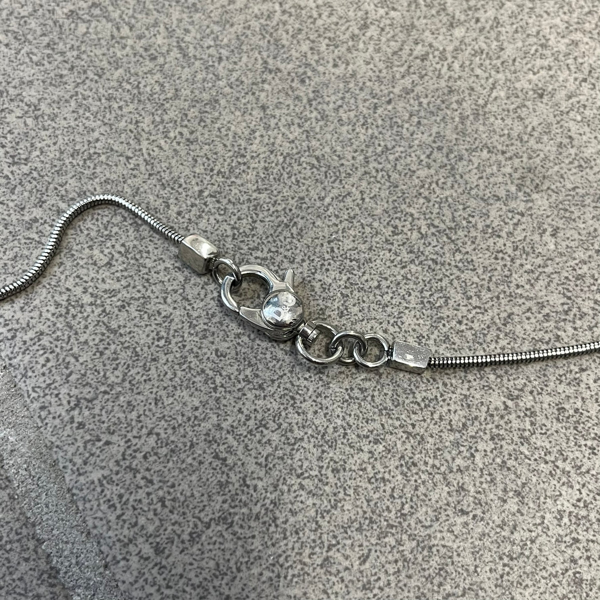 Dolce & Gabbana DJ1115 Circular Necklace is a stainless steel necklace with a circular pendant featuring a raised D&G logo in the center. A thin chain wraps around the pendant outline, held in place by small square clips at four points. It comes with a dust bag.