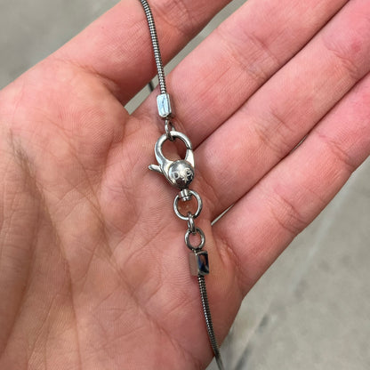 Dolce & Gabbana DJ1115 Circular Necklace is a stainless steel necklace with a circular pendant featuring a raised D&G logo in the center. A thin chain wraps around the pendant outline, held in place by small square clips at four points. It comes with a dust bag.