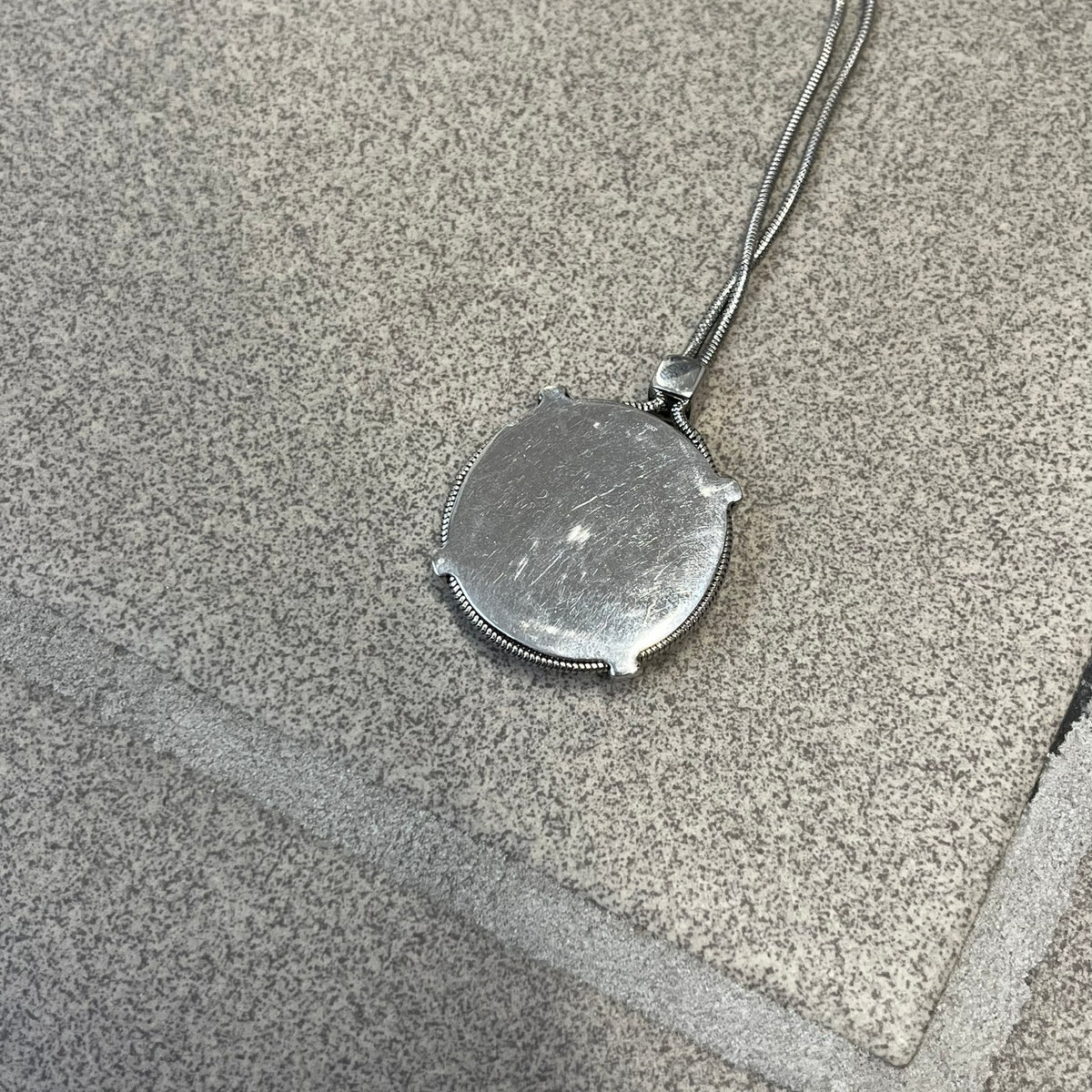 Dolce & Gabbana DJ1115 Circular Necklace is a stainless steel necklace with a circular pendant featuring a raised D&G logo in the center. A thin chain wraps around the pendant outline, held in place by small square clips at four points. It comes with a dust bag.
