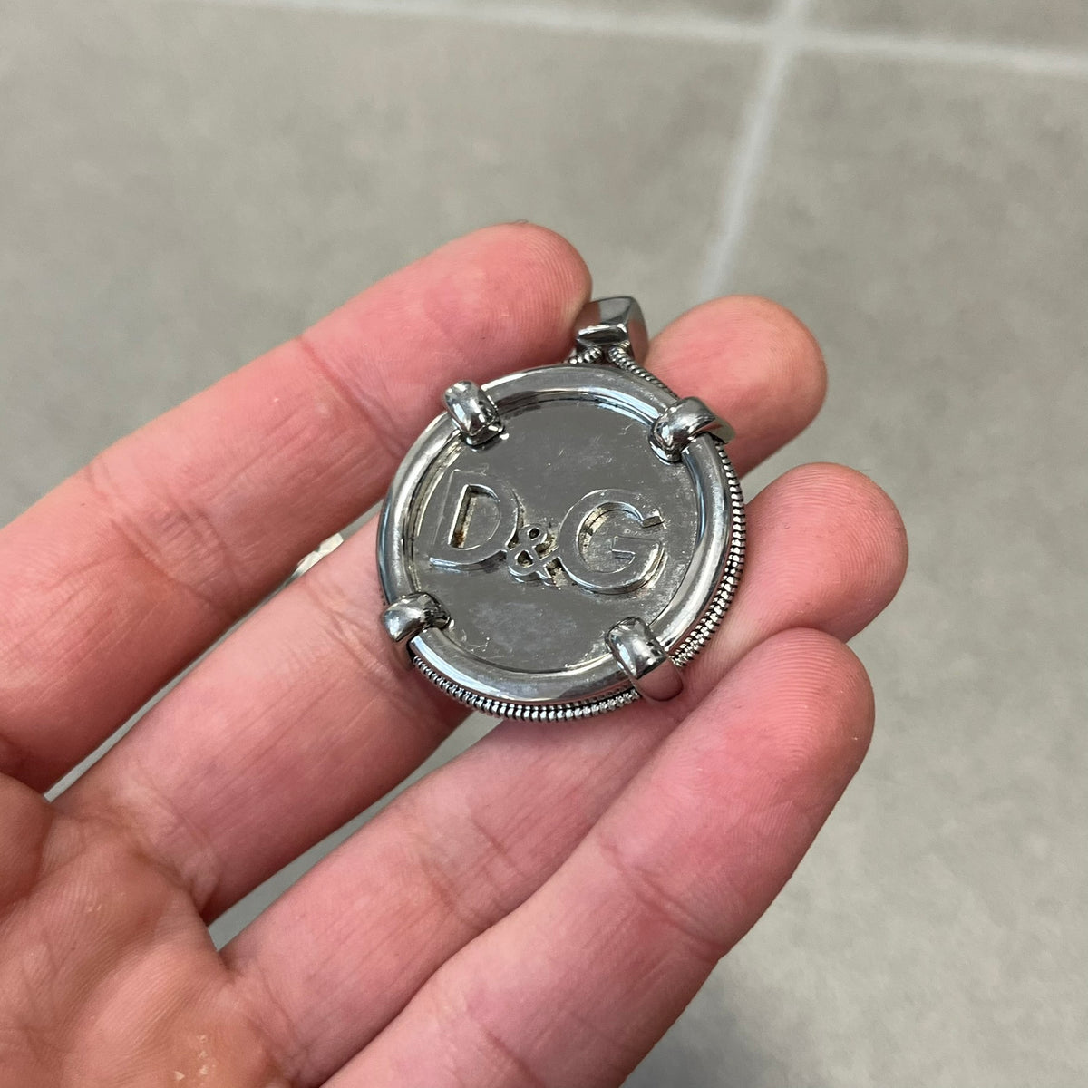 Dolce & Gabbana DJ1115 Circular Necklace is a stainless steel necklace with a circular pendant featuring a raised D&G logo in the center. A thin chain wraps around the pendant outline, held in place by small square clips at four points. It comes with a dust bag.