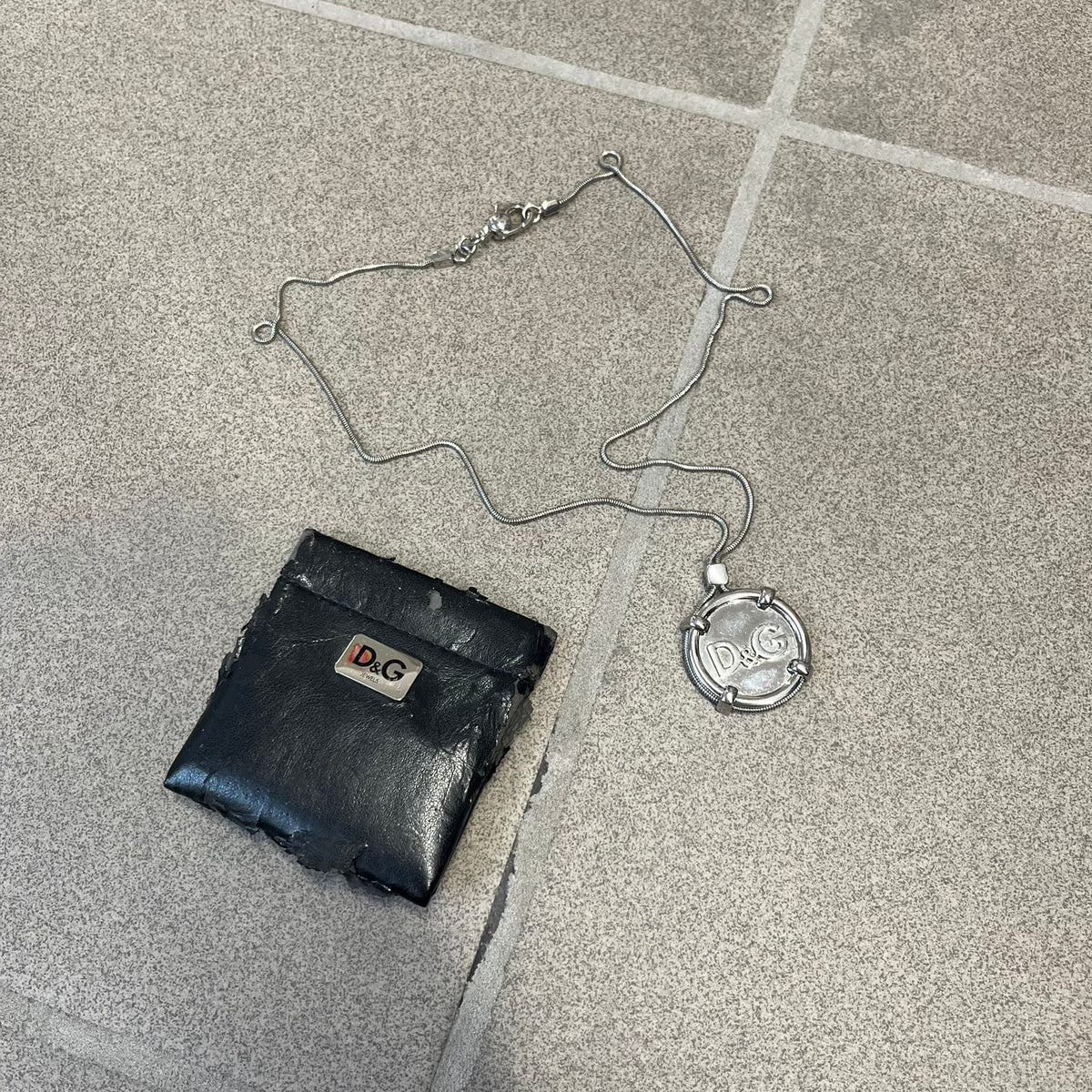 Dolce & Gabbana DJ1115 Circular Necklace is a stainless steel necklace with a circular pendant featuring a raised D&G logo in the center. A thin chain wraps around the pendant outline, held in place by small square clips at four points. It comes with a dust bag.