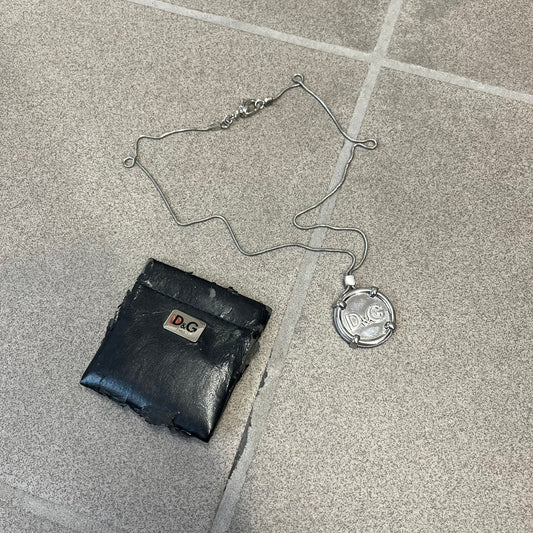 Dolce & Gabbana DJ1115 Circular Necklace is a stainless steel necklace with a circular pendant featuring a raised D&G logo in the center. A thin chain wraps around the pendant outline, held in place by small square clips at four points. It comes with a dust bag.