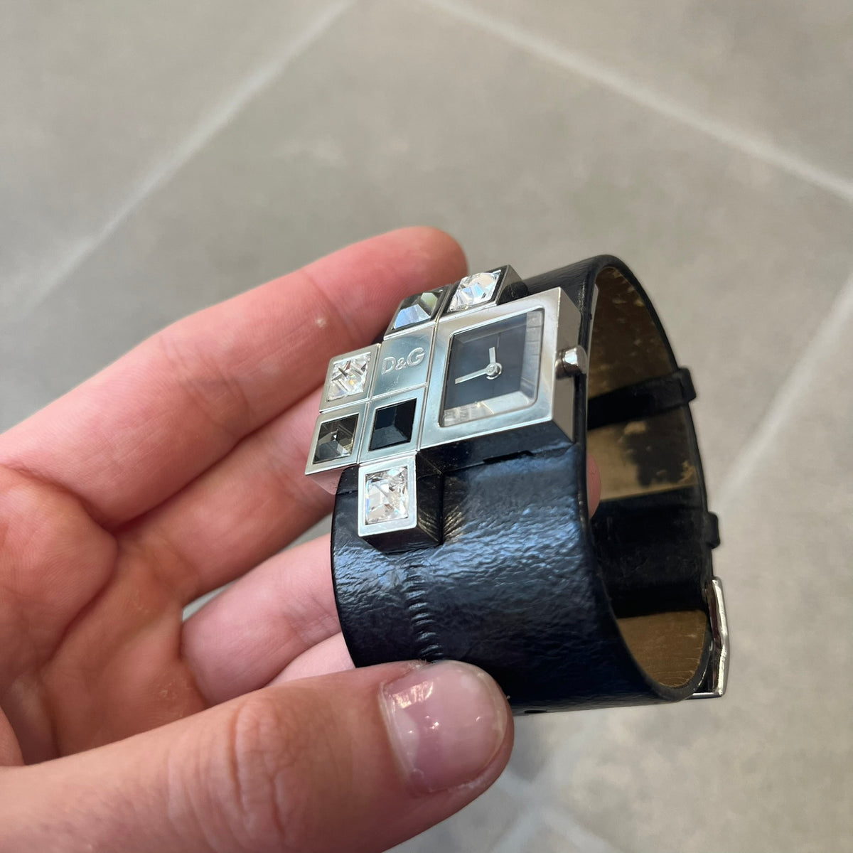 Dolce & Gabbana DW0174 Black Leather Analog Watch features a stainless steel dial composed of multiple squares, including smaller squares set with clear and black crystals, one bearing a D&G logo, and a larger square housing the watch hands. It is water resistant to 3 ATM.