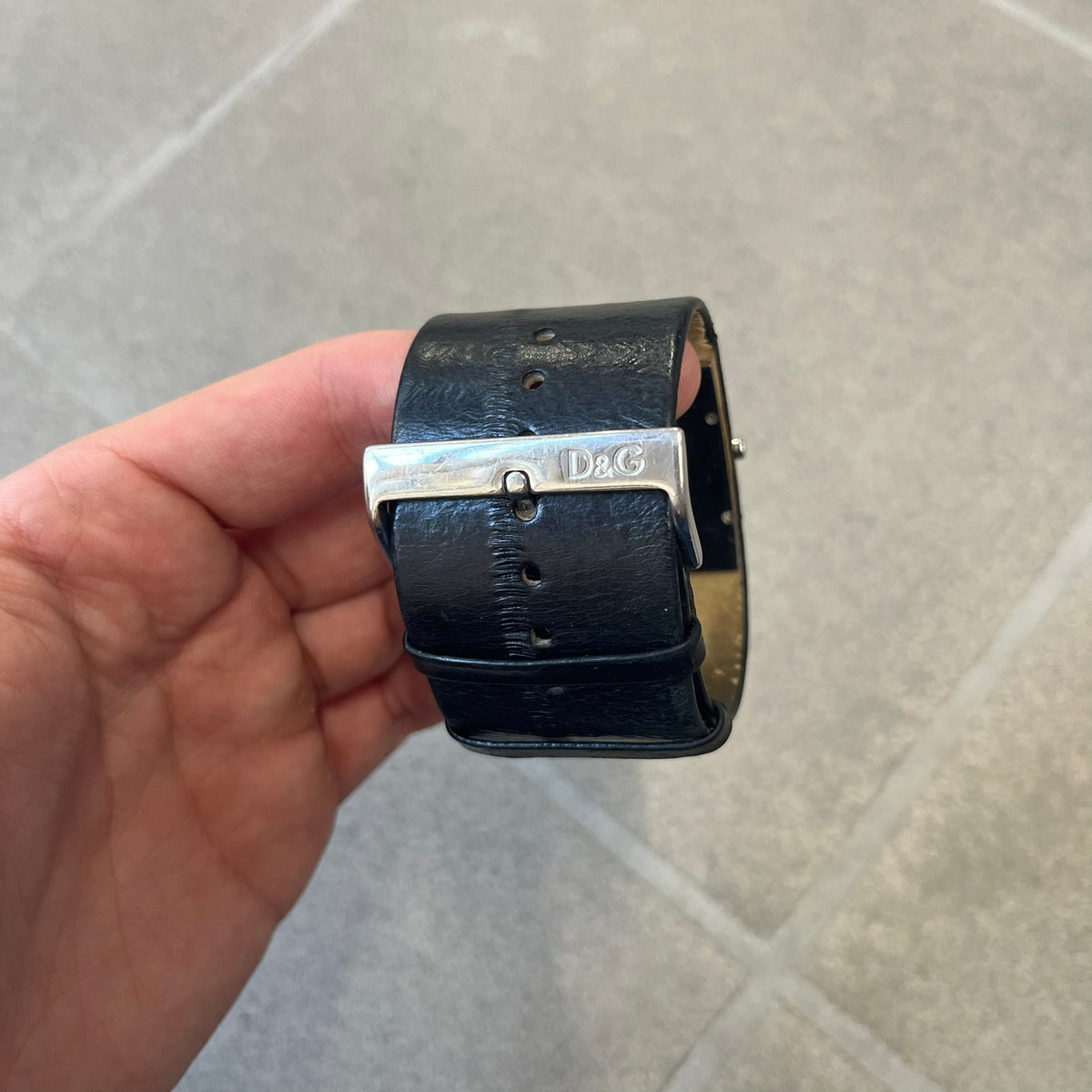 Dolce & Gabbana DW0174 Black Leather Analog Watch features a stainless steel dial composed of multiple squares, including smaller squares set with clear and black crystals, one bearing a D&G logo, and a larger square housing the watch hands. It is water resistant to 3 ATM.