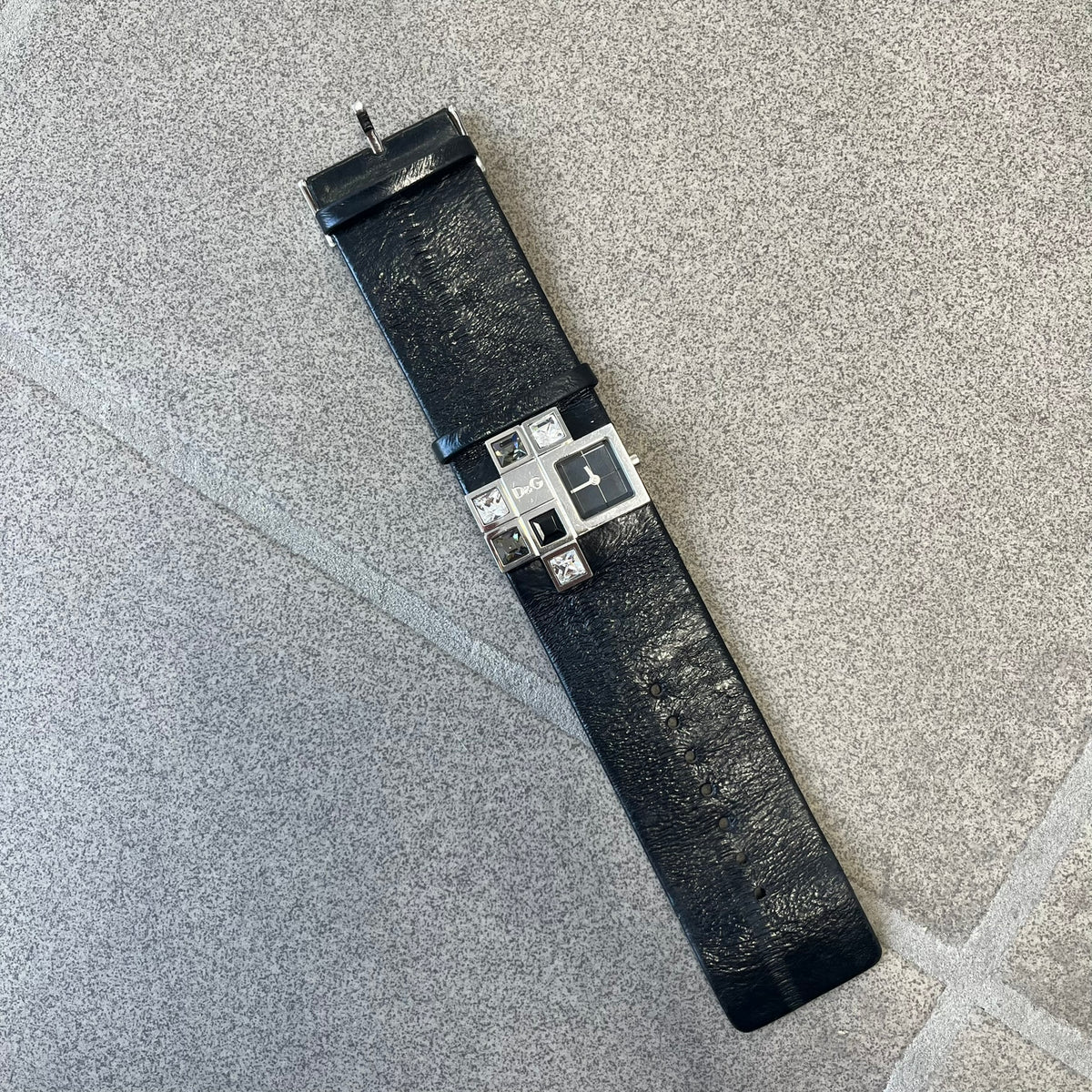 Dolce & Gabbana DW0174 Black Leather Analog Watch features a stainless steel dial composed of multiple squares, including smaller squares set with clear and black crystals, one bearing a D&G logo, and a larger square housing the watch hands. It is water resistant to 3 ATM.