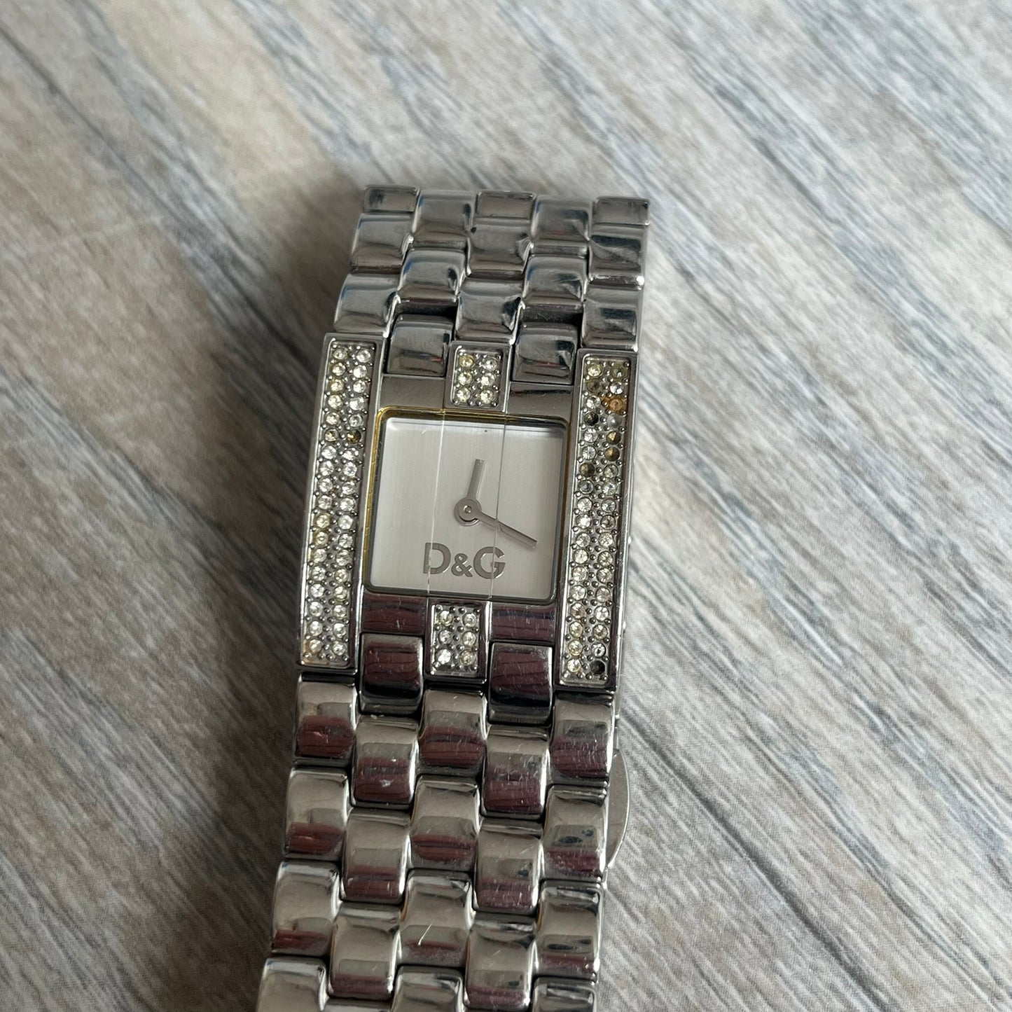 Dolce &amp; Gabbana Rhinestones Analog Watch is a stainless steel quartz watch featuring a square white dial with silver D&amp;G logo and silver hands. The case and bracelet form a continuous design with the face matching the width of the bracelet, and selected areas are set with crystal accents. It includes a dual side-clasp closure, is water resistant to 3 ATM, with removable links.