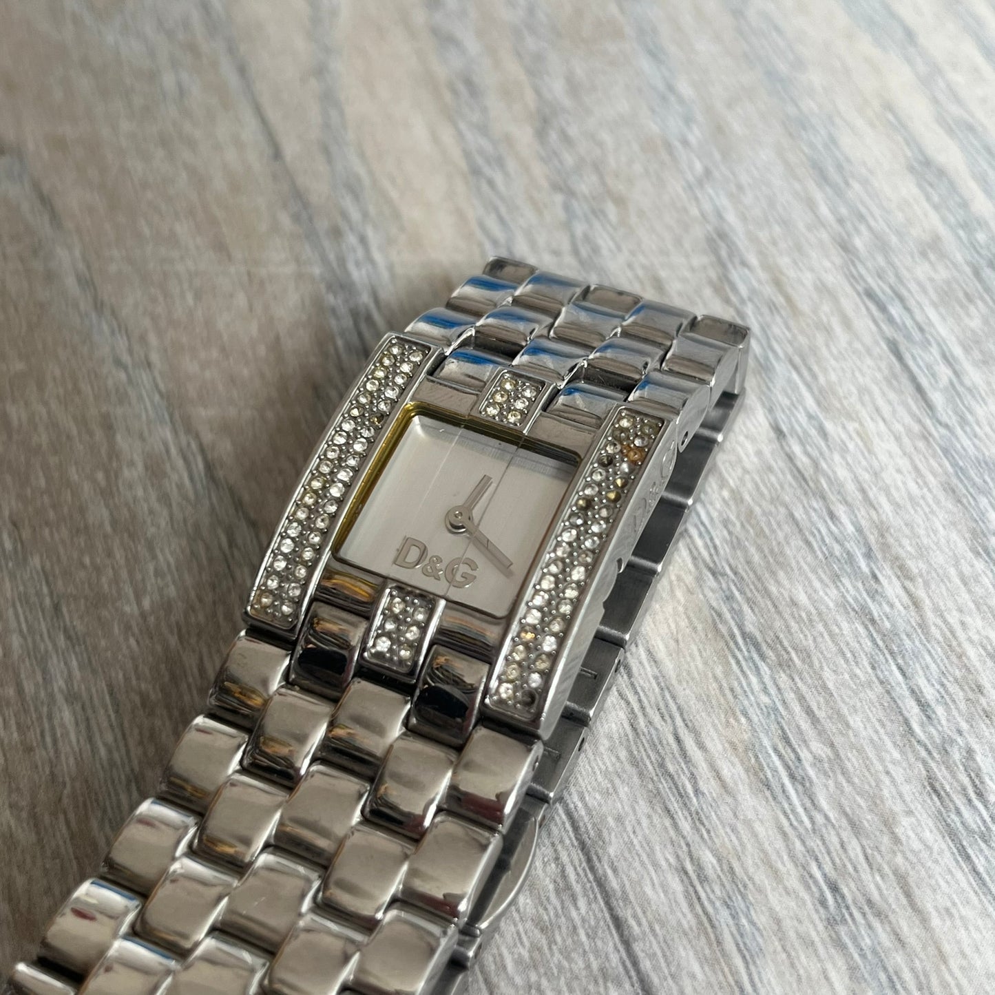Dolce &amp; Gabbana Rhinestones Analog Watch is a stainless steel quartz watch featuring a square white dial with silver D&amp;G logo and silver hands. The case and bracelet form a continuous design with the face matching the width of the bracelet, and selected areas are set with crystal accents. It includes a dual side-clasp closure, is water resistant to 3 ATM, with removable links.