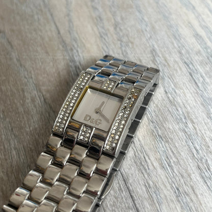 Dolce &amp; Gabbana Rhinestones Analog Watch is a stainless steel quartz watch featuring a square white dial with silver D&amp;G logo and silver hands. The case and bracelet form a continuous design with the face matching the width of the bracelet, and selected areas are set with crystal accents. It includes a dual side-clasp closure, is water resistant to 3 ATM, with removable links.