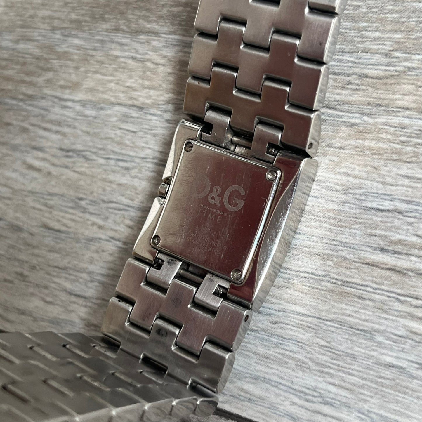 Dolce &amp; Gabbana Rhinestones Analog Watch is a stainless steel quartz watch featuring a square white dial with silver D&amp;G logo and silver hands. The case and bracelet form a continuous design with the face matching the width of the bracelet, and selected areas are set with crystal accents. It includes a dual side-clasp closure, is water resistant to 3 ATM, with removable links.