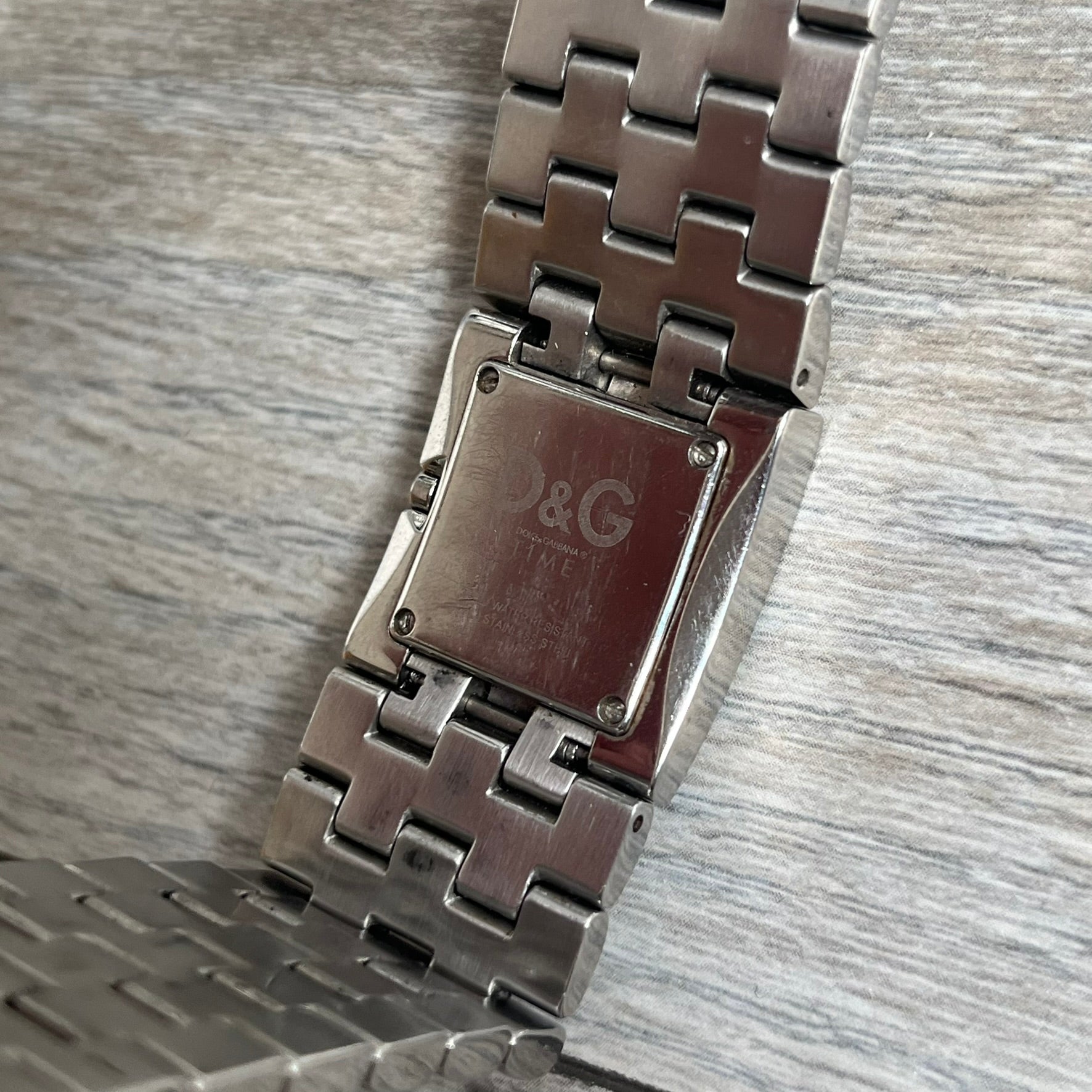 Dolce &amp; Gabbana Rhinestones Analog Watch is a stainless steel quartz watch featuring a square white dial with silver D&amp;G logo and silver hands. The case and bracelet form a continuous design with the face matching the width of the bracelet, and selected areas are set with crystal accents. It includes a dual side-clasp closure, is water resistant to 3 ATM, with removable links.