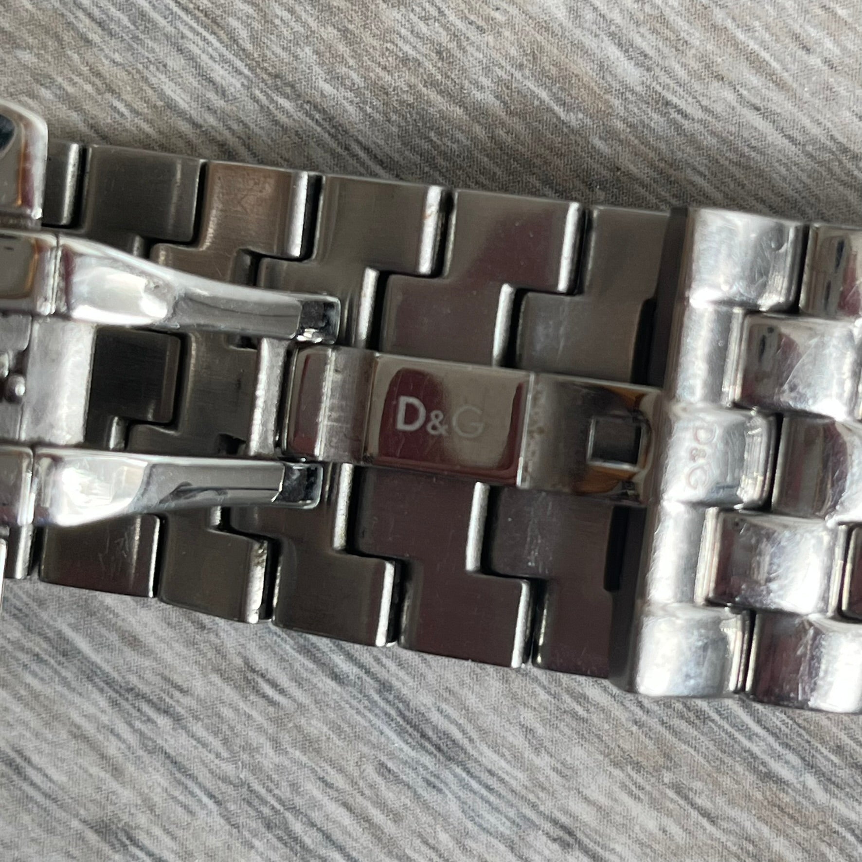 Dolce &amp; Gabbana Rhinestones Analog Watch is a stainless steel quartz watch featuring a square white dial with silver D&amp;G logo and silver hands. The case and bracelet form a continuous design with the face matching the width of the bracelet, and selected areas are set with crystal accents. It includes a dual side-clasp closure, is water resistant to 3 ATM, with removable links.