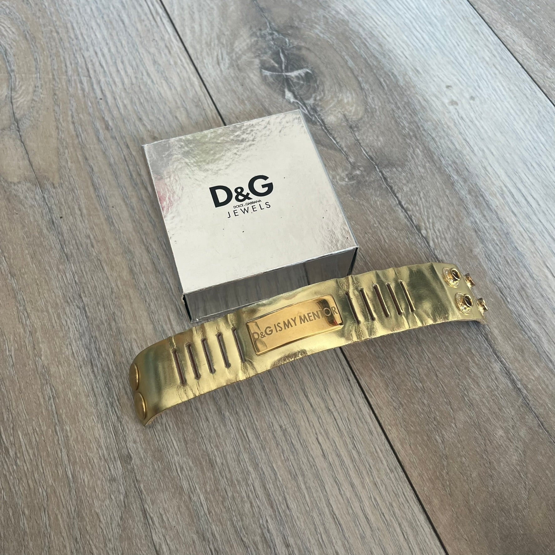 Dolce & Gabbana “D&G IS MY MENTOR” Bracelet is a gold leather bracelet with a glossy finish. The bracelet features line cutout detailing throughout and fastens with two snap-button closures on the back. A gold-tone metal plaque on the front is engraved with “D&G IS MY MENTOR.” The bracelet comes with its original box.
