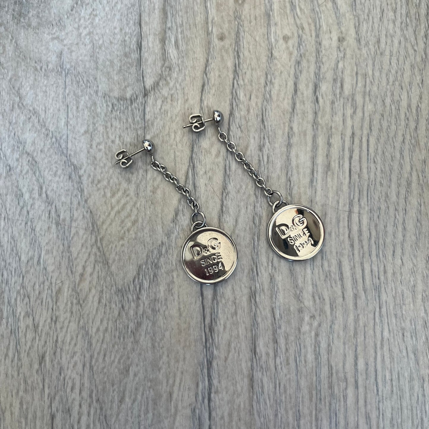 Dolce & Gabbana “D&G Since 1994” Earrings are a pair of 2000s silver-tone hanging earrings. Each earring features a short chain with a round pendant engraved with “D&G Since 1994.” The earrings have a polished metal finish and matching circular plaques.
