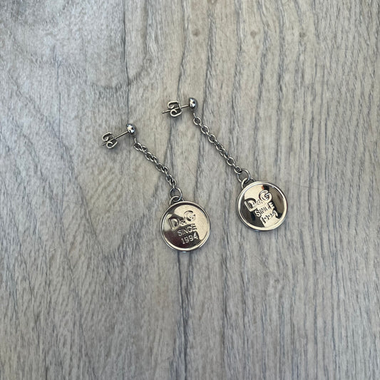 Dolce & Gabbana “D&G Since 1994” Earrings are a pair of 2000s silver-tone hanging earrings. Each earring features a short chain with a round pendant engraved with “D&G Since 1994.” The earrings have a polished metal finish and matching circular plaques.

